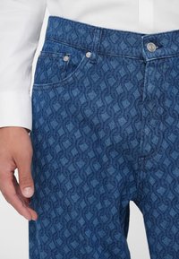 Blauwe denim jeans met een gestructureerd patroon van in elkaar grijpende vormen. Voorzien van een knoopsluiting en details van zijzakken.