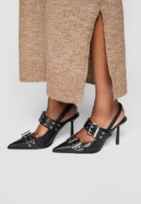 Črne usnjene pete s slingback oblikovanjem in koničastimi prsti, ki vključujejo srebrne zaponke in eleganten špicasti peto. Tekstura se zdi gladka in sijajna.