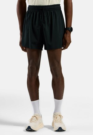 Schwarze Sportshorts aus leichtem Material, mit einem elastischen Bund und einer Kordel. Kombiniert mit weißen Kniestrümpfen und beigen Sneakers.