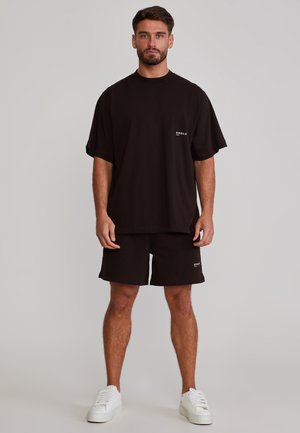 Férfi áll fekete túlméretezett pólóban és hozzá illő rövidnadrágban, fehér tornacipőben, egyszerű háttér előtt.