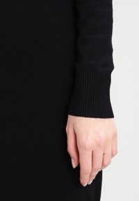 Czarna dzianinowa sukienka z detalami na mankietach w prążki. Tekstura jest gładka i dopasowana, sięgająca do nadgarstka z ciasnym wykończeniem.