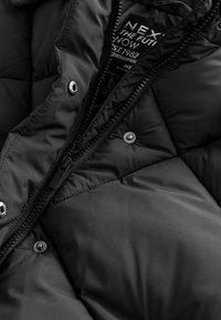 Veste matelassée noire avec fermeture éclair visible, boutons-pression métalliques et étiquette intérieure partiellement visible indiquant "NEX THE FUTURE NOW EST 1982."
