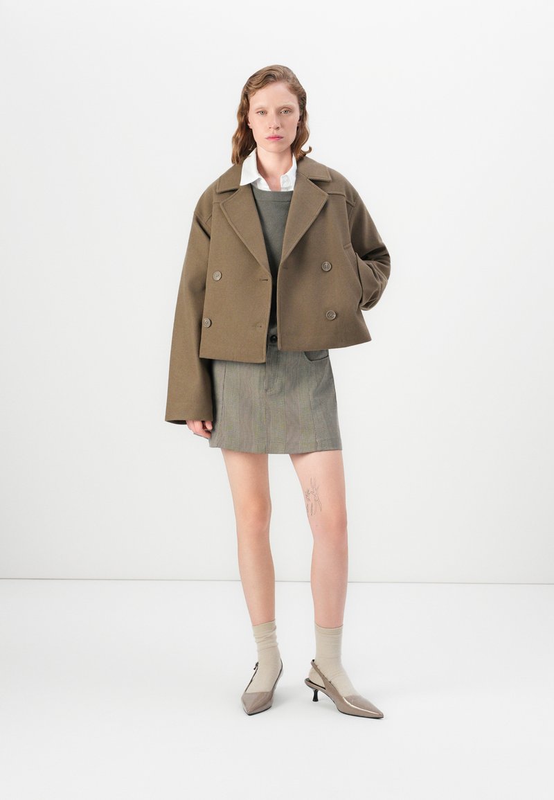 Veste marron croisée à double boutonnage sur un haut gris et une jupe mini à carreaux. Le modèle porte des chaussettes beiges arrivant au genou et des chaussures à talons pointues.