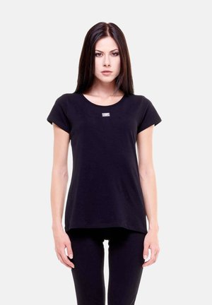 Giovane donna con lunghi capelli scuri che indossa una semplice maglietta nera a maniche corte e pantaloni neri, in piedi su uno sfondo bianco.