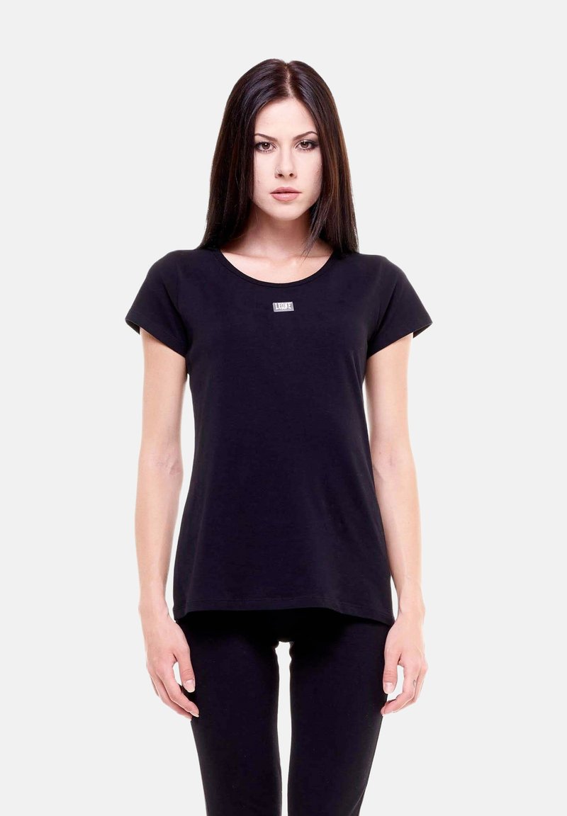 Jeune femme aux longs cheveux foncés, portant un t-shirt noir uni à manches courtes et un pantalon noir, debout devant un fond blanc.