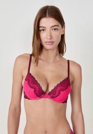 Femme à la peau claire et aux cheveux bruns portant un soutien-gorge en dentelle rose et bourgogne, se tenant devant un fond clair uniforme.