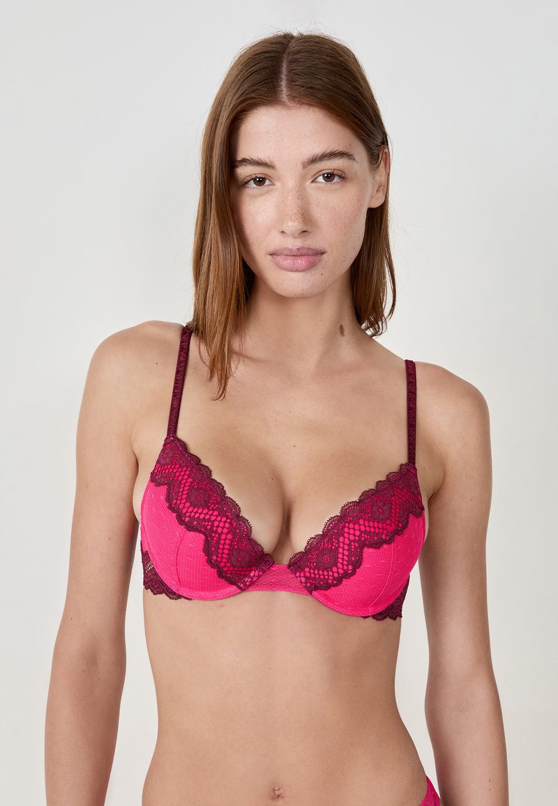 Vrouw met een lichte huid en bruin haar, gekleed in een roze en burgundy kanten bh, staand tegen een effen lichte achtergrond.