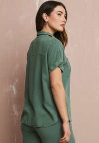 Camicia verde maniche corte con colletto, in tessuto morbido e maniche arrotolate. Presenta una vestibilità comoda e un orlo arrotondato per un aspetto informale.