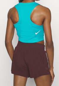 Top corto senza maniche turchese con design a racerback e logo Nike bianco, abbinato a pantaloni corti larghi color amaranto. Tessuto morbido, stile minimalista.