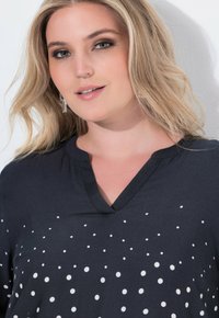 Top nero con scollo a V e pois bianchi di dimensioni variabili. Tessuto morbido e vestibilità rilassata, con un colletto pulito e dettagli delle cuciture sottili.
