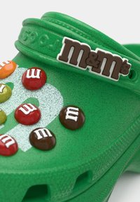 Green Crocs featuring a glossy upper, M&M's candy embellishments in various colors, and a white logo accent on the side. Textured finish.