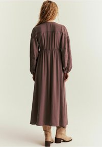 Długa, zwiewna sukienka w kolorze mauve z marszczoną talią, długimi puffowymi rękawami i haftem kwiatowym. Połączona z beżowymi kozaczkami sięgającymi za kolano na neutralnym tle.