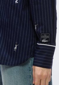 Chemise bleu marine à rayures verticales blanches, ornée de joueurs de tennis brodés et d'une étiquette noire sur la manche. Matière en coton, poignets à boutons.