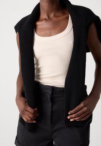 Cárdigan negro de punto con un frente abierto, tejido texturizado y hombros caídos, combinado con una camiseta sin mangas beige y pantalones cortos oscuros.