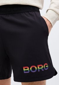 Μαύρα σορτς με πολύχρωμο λογότυπο "BORG" στο αριστερό πόδι, φορεμένα με ανοιχτόχρωμη μπλούζα με μακριά μανίκια· δεξί χέρι στην τσέπη.