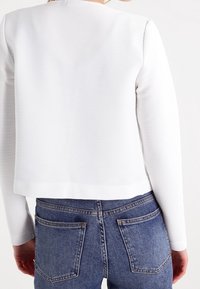 Chaqueta corta blanca con relieve y mangas largas. Presenta un cuello redondo y un dobladillo recto, combinada con pantalones vaqueros de talle alto.