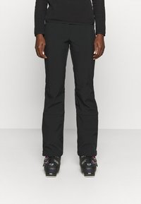 Schwarze Skihosen aus wasserabweisendem Stoff, slim fit, mit Reißverschlusstaschen und verstärkten Säumen, kombiniert mit schwarzen Skischuhen.