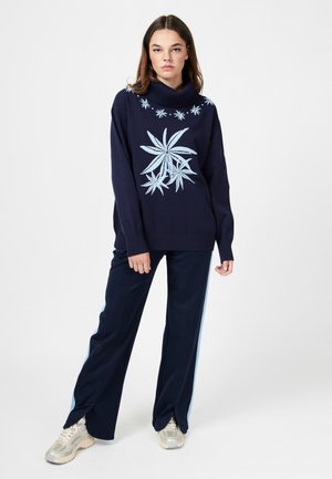 Granatowy sweter z jasnoniebieskim haftem w kwiaty, wysoki kołnierz i luźny krój. Połączony z granatowymi spodniami z jasnoniebieskimi bocznymi paskami i trampkami.