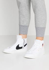 Valkoiset korkeat tennarit mustilla yksityiskohdilla ja oranssilla yksityiskohdalla, joissa on vetoketju sivussa. Valmistettu kankaasta, jossa on tekstuuripinta.