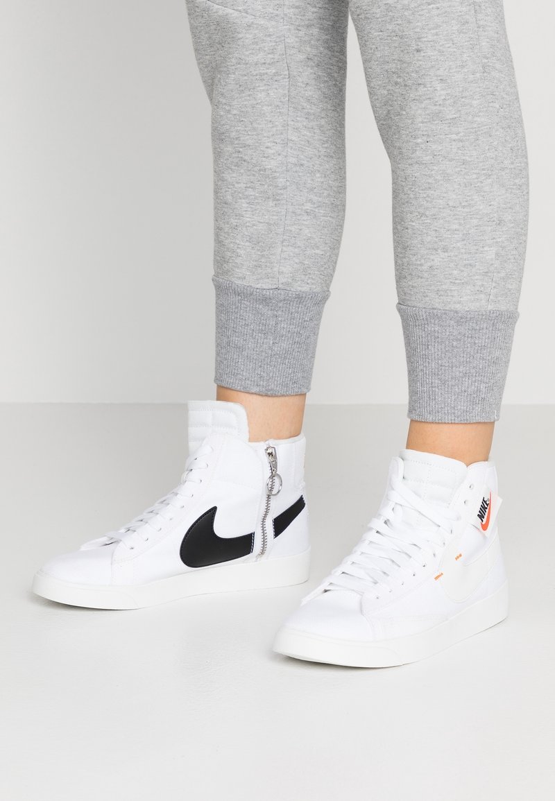 Valkoiset korkeat tennarit mustilla yksityiskohdilla ja oranssilla yksityiskohdalla, joissa on vetoketju sivussa. Valmistettu kankaasta, jossa on tekstuuripinta.