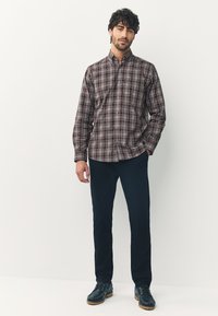 Chemise à carreaux dans des tons marron et beige, col boutonné, manches longues, associée à un pantalon foncé et des bottes. Texture du tissu lisse.