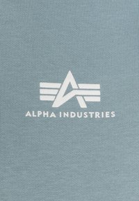 Világoskék pamut anyag, fehér logóval, rajta az "ALPHA INDUSTRIES" felirattal és geometriai díszítésekkel. Sima tapintású, bordázott kialakítású.