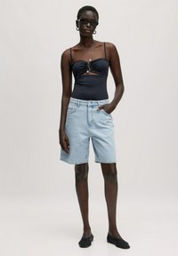 Zwarte zwemkleding met geplooide details en zilveren hardware, gecombineerd met lichtblauwe spijkerbroekshorts en zwarte instapschoenen. Zonnebril maakt de look compleet.