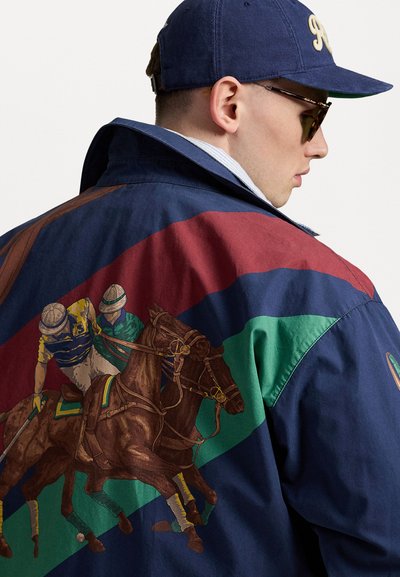 Veste bleu marine avec une broderie colorée de cheval et cavalier, ornée de rayures diagonales vertes et bordeaux et d'un tissu texturé.