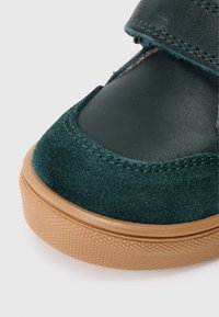 Grüner Lederschuh mit Wildleder-Zehenkappe und tannfarbener Gummisohle. Verfügt über eine abgerundete Form und zwei Klettverschlüsse zur Sicherung. Texturierte Sohle.