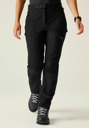 Personne portant un pantalon de plein air noir avec des poches zippées, une ceinture noire et des chaussures de randonnée sombres avec des lacets gris clair marchant vers l'avant.