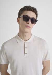 Bleke beige polo met korte mouwen en een drieknoopsluiting. Zachte textuur, met subtiele logodetail op de linkerborst. Zwarte zonnebril.