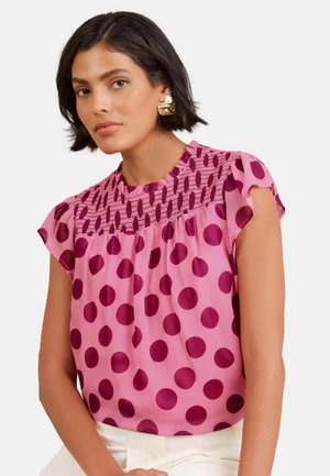 Vrouw met kort zwart haar die grote gouden oorbellen draagt en een roze blouse met donkerroze stippen en een geplooide halslijn.