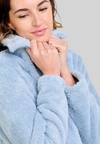 Chaqueta de tela suave y esponjosa de color azul claro con un cuello alto y puños enrollados. La textura parece suave y cálida. El modelo está agarrando el cuello.
