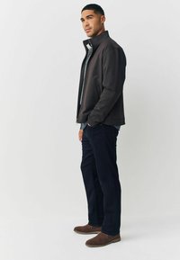 Pánská tmavě hnědá bunda s vysokým límcem, doplněná vzorovanou košilí, tmavými džínami a hnědými semišovými botami. Hladká textura a volný střih.