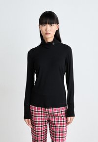 Schwarzes Langarmoberteil mit umgeschlagenem Kragen und dekorativem Verschluss. Weiche Textur mit gerippten Bündchen. Kombiniert mit einer Hose im pinken und schwarzen Hahnentrittmuster.