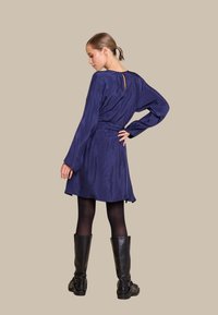 Marineblå kjole med lange ermer, strukturert stoff, belte i midjen og detalj med nøkkelhull bak, paret med svarte knelengde støvler og svarte strømpebukser.