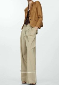 Hnědý strukturovaný blejzr, béžové plisované kalhoty s širokými nohavicemi a bílým prošíváním, vrstvené přes tmavý top. Hladká tkanina s dobře padnoucím střihem.