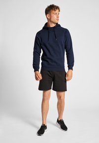 Marineblaues Kapuzenshirt mit langen Ärmeln und einer Kängurutasche, kombiniert mit schwarzen Shorts und schwarzen Sportschuhen. Einfaches, sportliches Design.