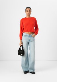 Rød kort genser med teksturert stoff, lyseblå vide jeans og en liten svart håndveske med hvit aksent. Svart sko fullfører antrekket.