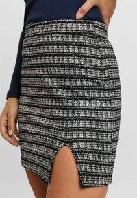 Kvinna iklädd svart och vit texturerad minikjol med frontslits, parat med långärmad marinblå tröja, hand som vilar på midjan.