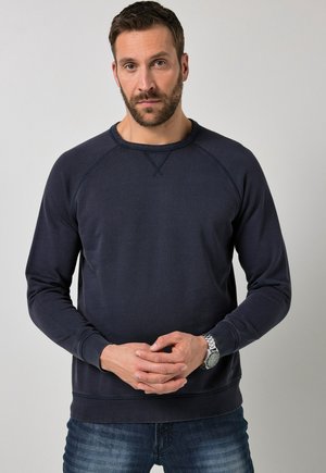 Homme aux cheveux courts et foncés et à la barbe, portant un sweat-shirt bleu marine et un jean, debout les mains jointes et portant une montre-bracelet argentée.