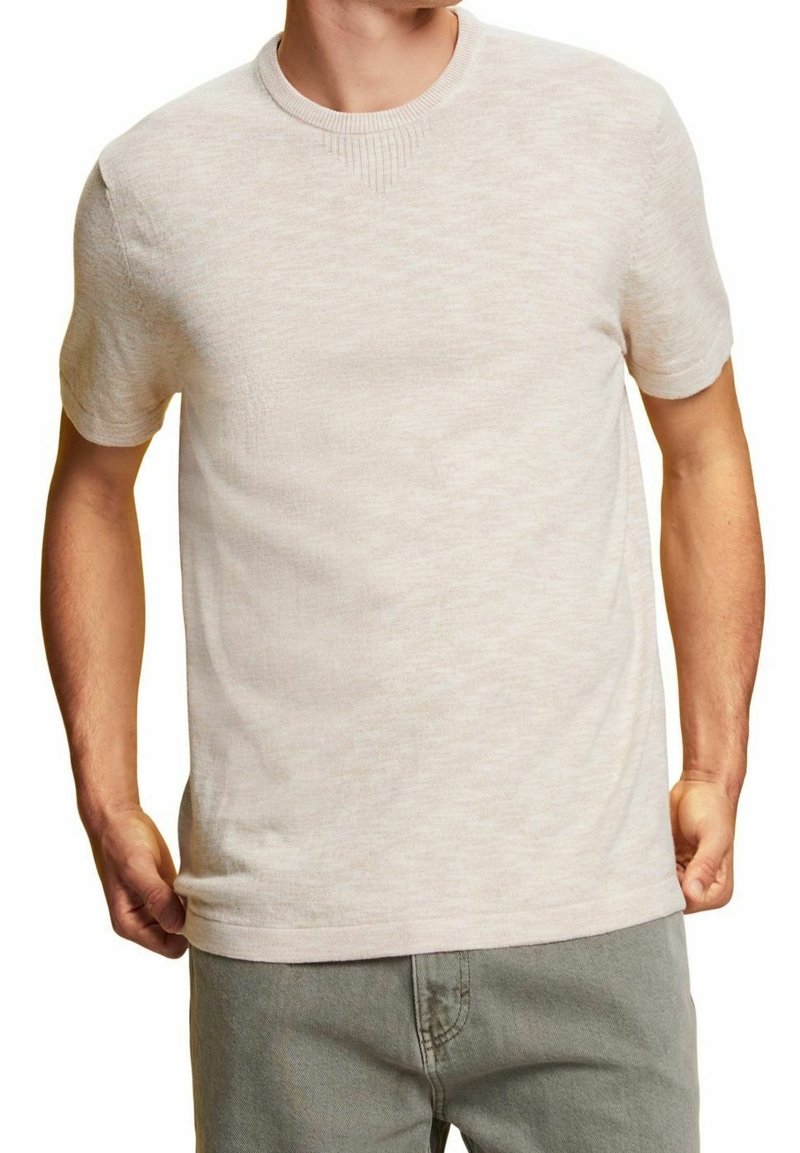 Chemise beige clair à manches courtes en tissu doux et texturé, avec un col rond et un bord-côtes subtil au niveau du col, cintrée près du corps.