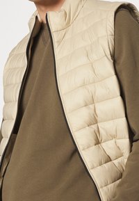 Gilet imbottito beige con un design trapuntato, colletto alto e dettagli della cerniera nera, indossato sopra una maglietta a maniche lunghe verde oliva.