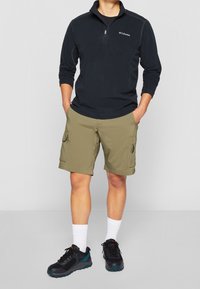 Musta vetoketjullinen fleece-pullove, jossa on pitkät hihat, yhdistettynä oliivinvihreisiin cargo-shorteihin, joissa on sivutaskut, ja mustiin lenkkikenkiin, joissa on sinisiä yksityiskohtia.