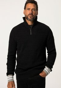 Maglione a coste nere con colletto con zip, dotato di polsini a strisce grigie e bianche, indossato da una persona con un'espressione neutra.