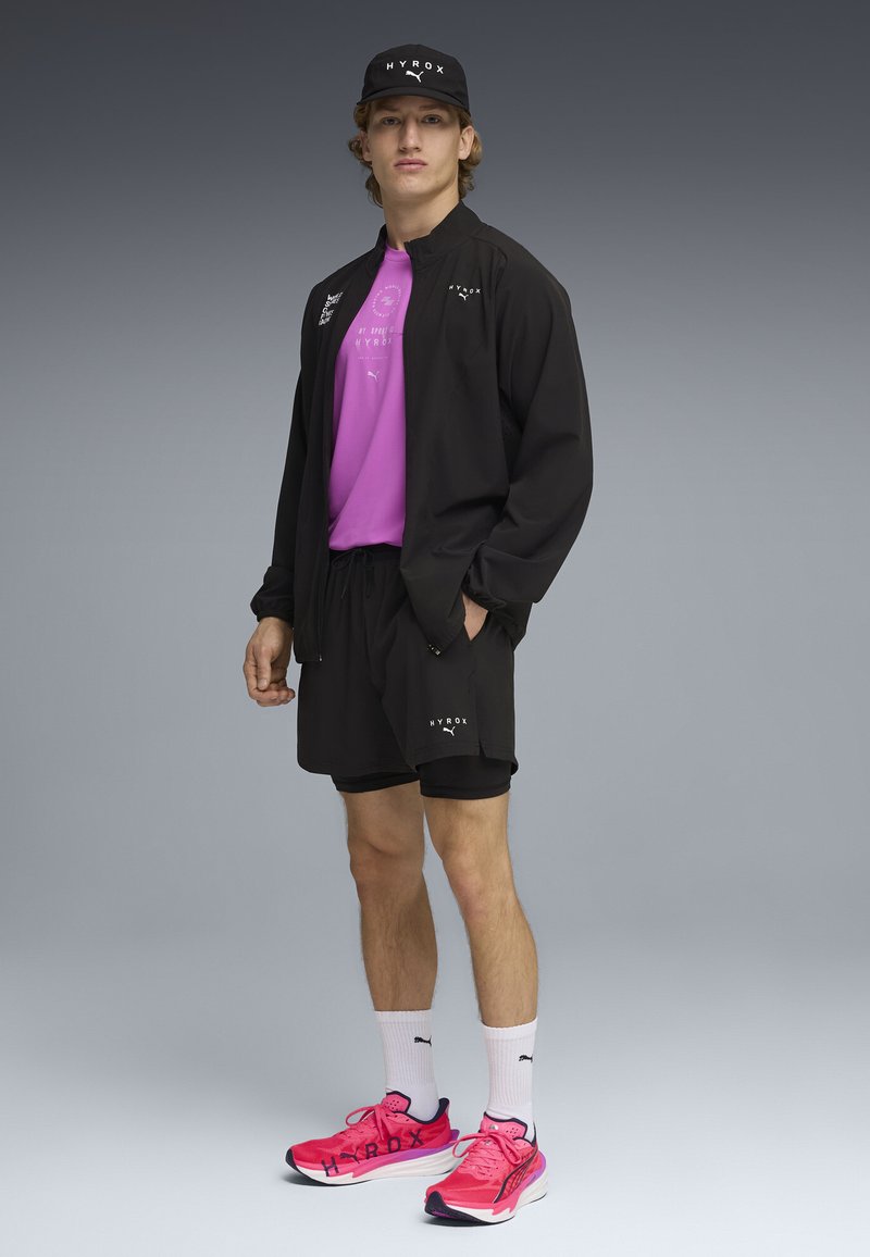 Jeune homme debout portant une casquette HYROX noire, une veste noire, un short noir, un t-shirt violet, des chaussettes blanches Puma et des chaussures de course rose vif.
