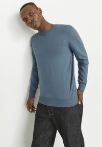 Helles blaues Crew-Neck-Pullover aus weichem Material, mit gerippten Bündchen und Saum, kombiniert mit dunklen Denim-Jeans mit farbigem Nähten.