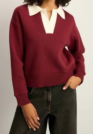 Femme portant un pull bordeaux à manches longues avec un col blanc, associé à un jean noir taille haute, les mains détendues près des poches.