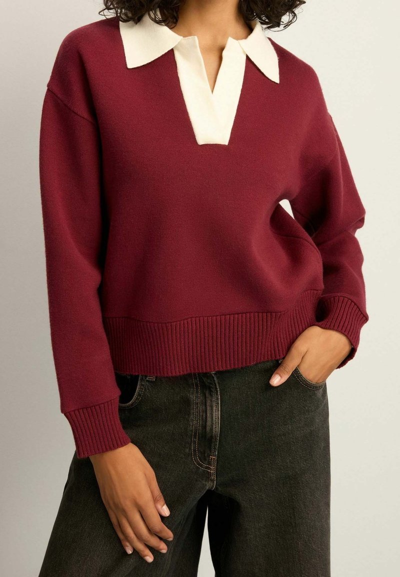 Femme portant un pull bordeaux à manches longues avec un col blanc, associé à un jean noir taille haute, les mains détendues près des poches.