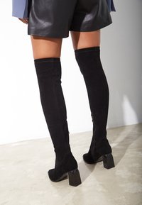 Botas negras de ante hasta la rodilla con tacón grueso. Presentan una textura suave, detalle de costura vertical y cremallera lateral para facilitar su uso.